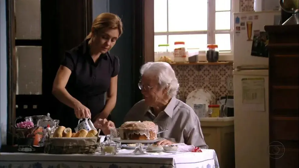 Uma mulher jovem serve café para uma senhora idosa sentada à mesa em uma cozinha aconchegante, onde há um bolo caseiro, pães e utensílios dispostos, com luz suave entrando pela janela ao fundo.