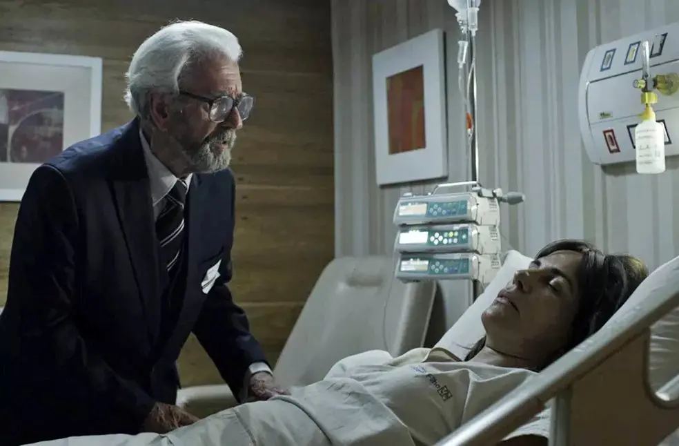 Um homem idoso de terno inclina-se ao lado de uma mulher deitada em um leito hospitalar, enquanto equipamentos médicos e um soro estão posicionados ao fundo do quarto.
