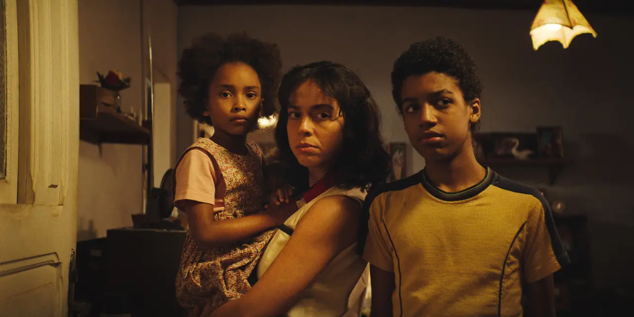 Três pessoas estão em um ambiente interno com iluminação quente. Uma pessoa adulta está ao centro, segurando uma criança pequena no colo, enquanto outra pessoa jovem está posicionada ao lado. As três estão voltadas para a câmera. O espaço ao fundo inclui móveis simples, uma prateleira com objetos e quadros, e uma luminária pendente no canto superior direito.