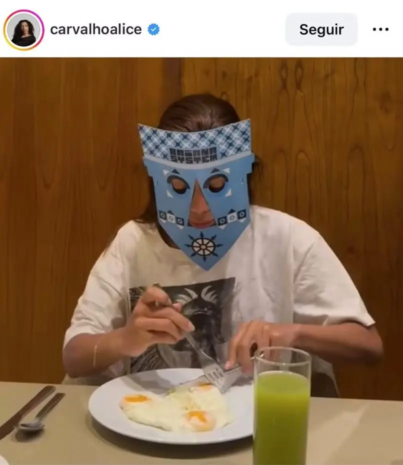 Pessoa sentada à mesa, usando uma máscara de papel azul com recortes geométricos que cobrem o rosto. A pessoa veste uma camiseta larga e segura garfo e faca, cortando dois ovos fritos em um prato branco. À direita do prato há um copo transparente com uma bebida verde.