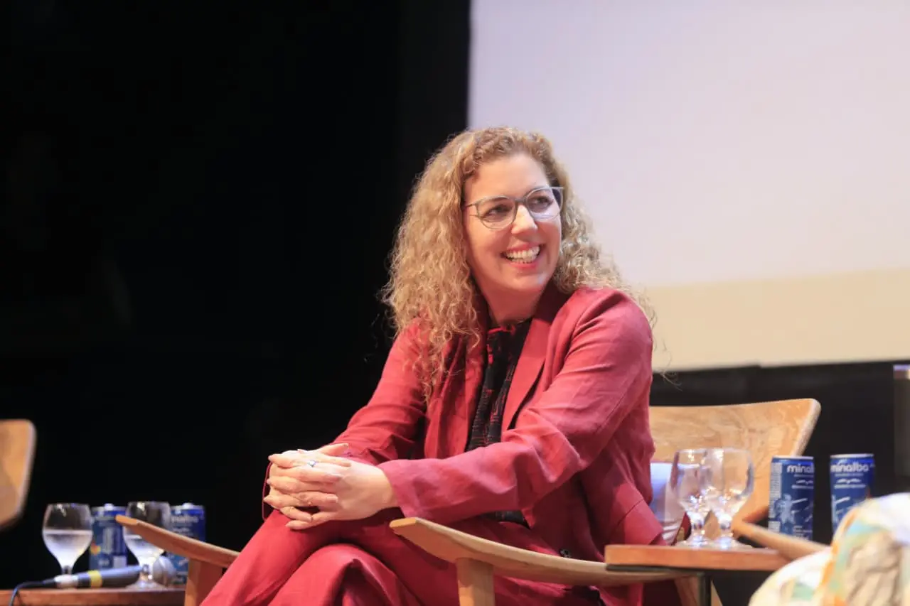 Ticiana Rolim. Mulher de blazer vermelho sentada em poltrona durante evento, sorrindo em palco com iluminação focada.