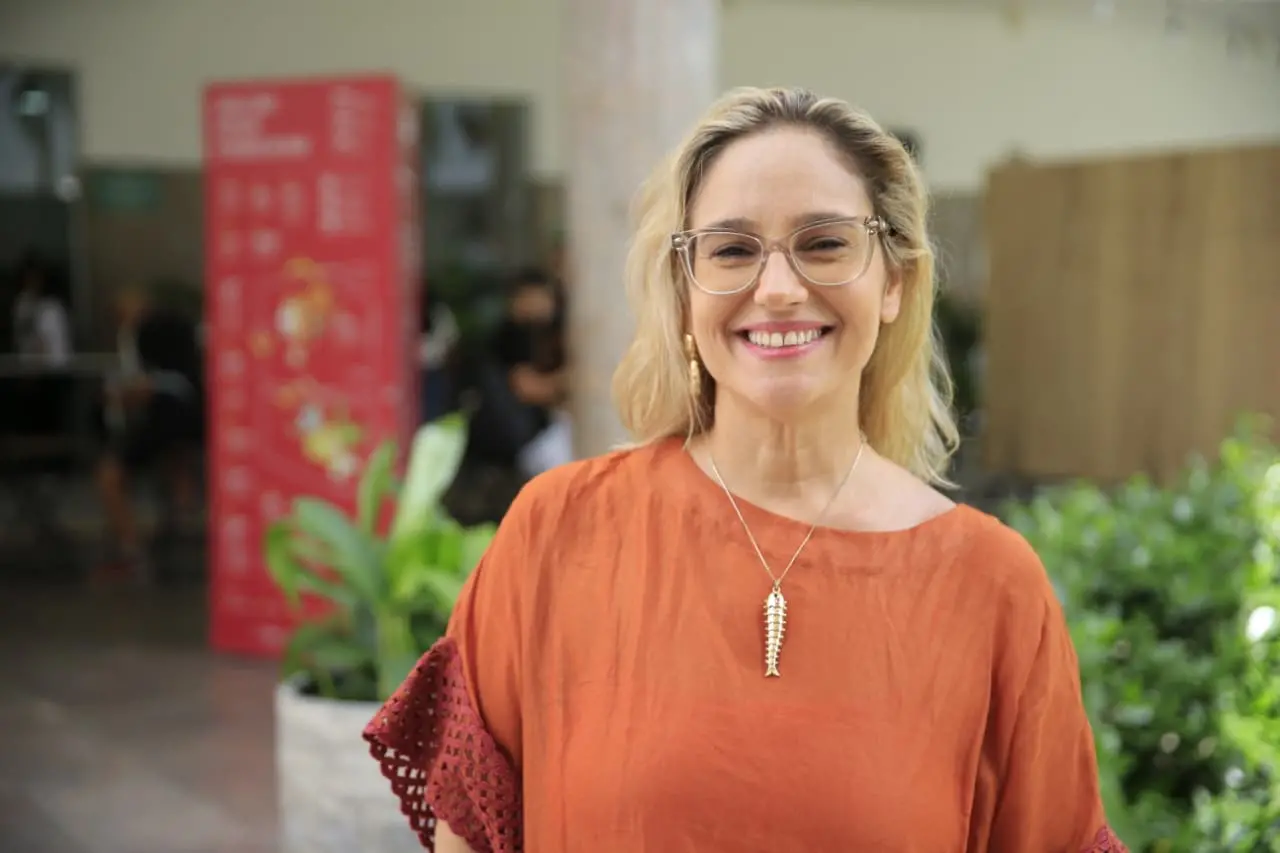 Lídice Berman. Mulher de óculos e blusa laranja sorri para a câmera em espaço interno com plantas e pessoas ao fundo.