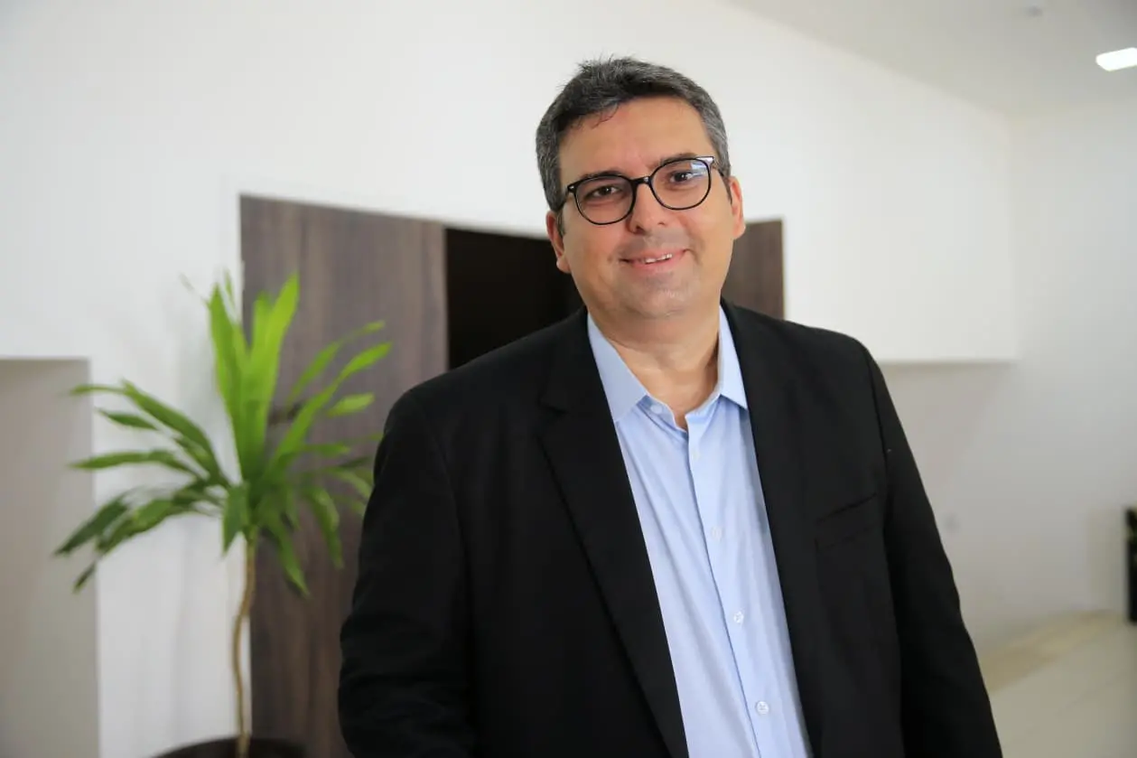 secretário executivo da SDE, Rennys Frota. Homem de terno e óculos sorri para a câmera em ambiente corporativo com planta ao fundo.