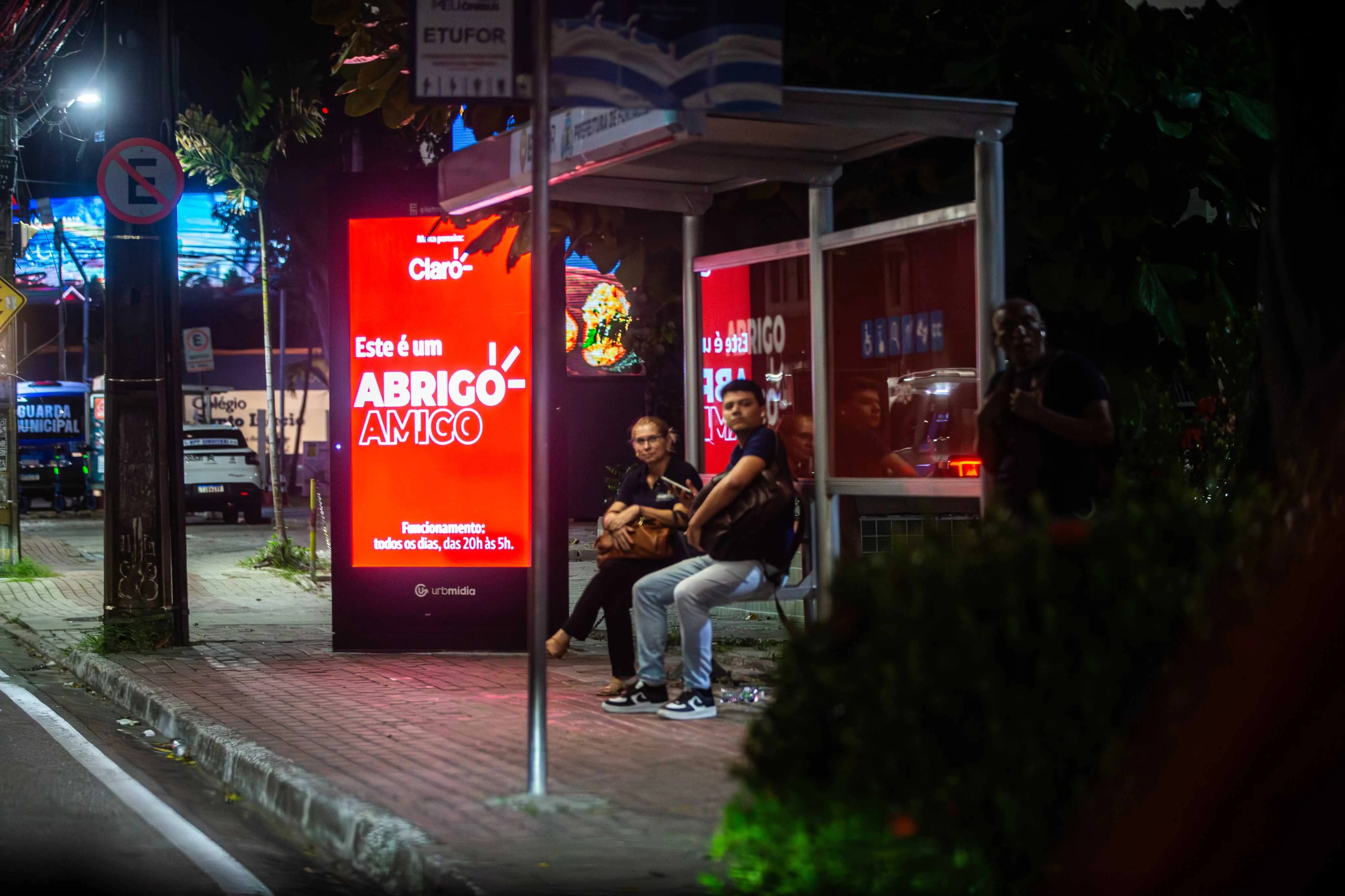 Parada de ônibus iluminada à noite com totem digital vermelho exibindo Este é um Abrigo Amigo. Duas pessoas esperam sentadas sob a cobertura, enquanto um carro da Guarda Municipal aparece ao fundo.