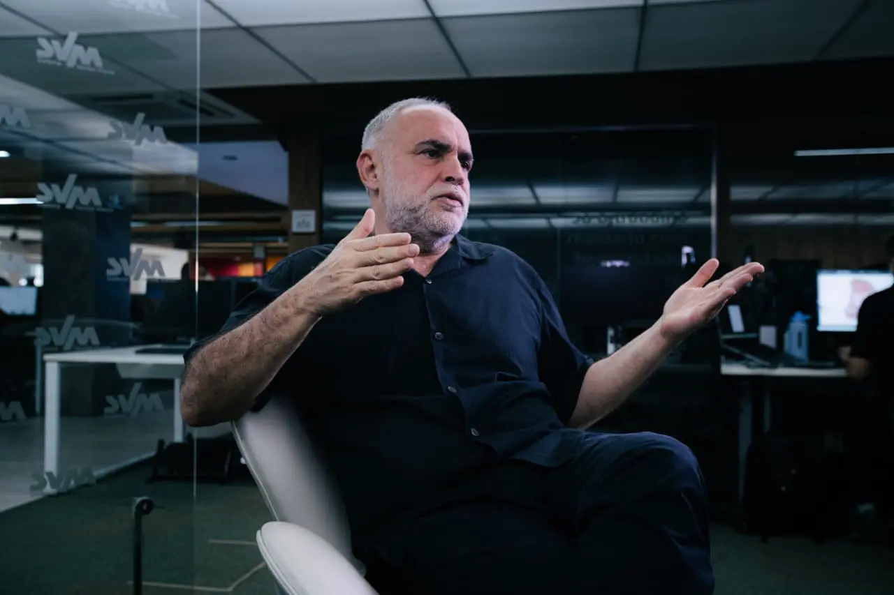 O cineasta Karim Aïnouz gesticula com as mãos abertas enquanto fala, sentado em uma poltrona branca em um ambiente de escritório contemporâneo. Ele veste uma camisa preta e está posicionado diante de uma divisória de vidro transparente com logomarcas, sob uma iluminação de teto suave.