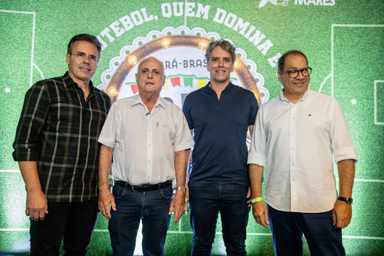 Quatro pessoas em pé, lado a lado, diante de um painel temático de futebol verde, com marcações de campo e um arco iluminado ao fundo.