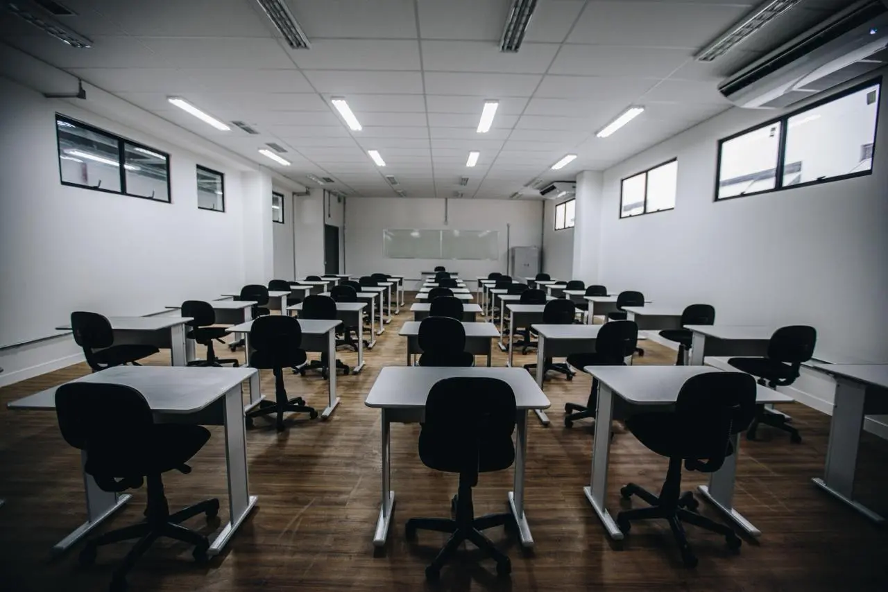 fotografia em ângulo aberto de uma sala de aula moderna, organizada e vazia. A perspectiva parte do fundo da sala, olhando em direção à lousa branca ao fundo.