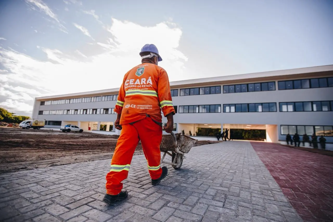 fotografia em ângulo de contra-plano mostra um trabalhador da construção civil de costas, caminhando por um caminho pavimentado de pedras em direção a um grande e moderno prédio escolar. Ele veste um macacão laranja vibrante com faixas refletivas amarelas e um capacete de segurança azul. Ele empurra um carrinho de mão de metal em direção ao prédio.
