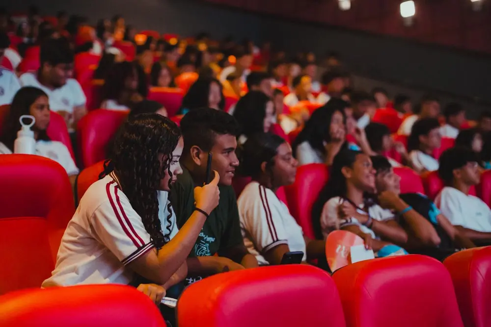 Estudantes ocupam as poltronas vermelhas de um cinema lotado, com o foco em uma jovem de cabelos cacheados que observa atentamente a tela enquanto segura um celular. Ao seu redor, outros jovens em uniformes escolares conversam e aguardam o início da sessão em um ambiente de luz suave e tons quentes.