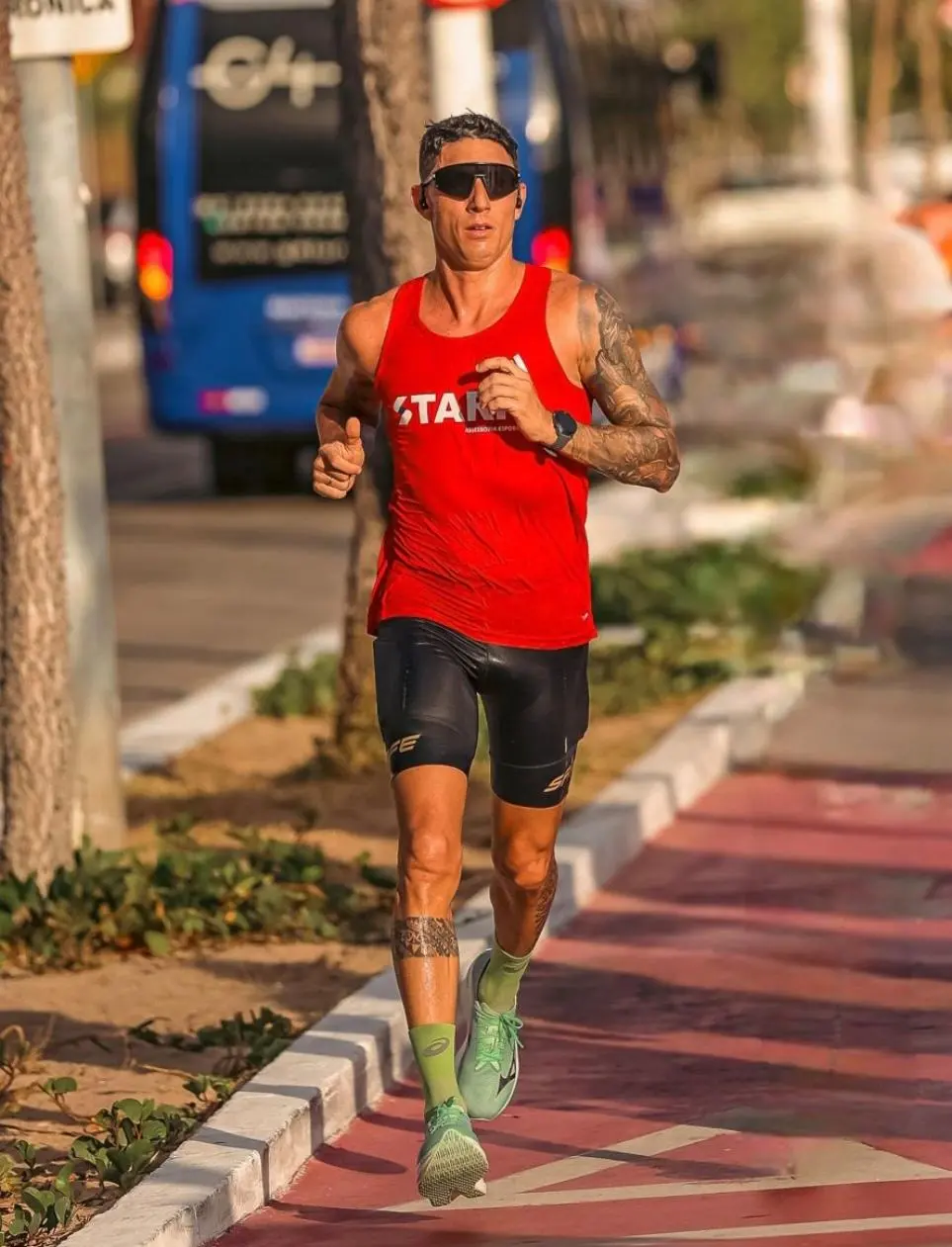 Pessoa correndo em uma ciclovia urbana, usando regata vermelha, bermuda preta e tênis verdes, com ônibus e árvores ao fundo em um dia ensolarado.