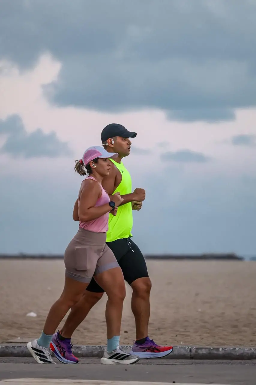 Duas pessoas correndo lado a lado na orla, usando roupas esportivas, em uma pista próxima à areia da praia sob céu nublado.