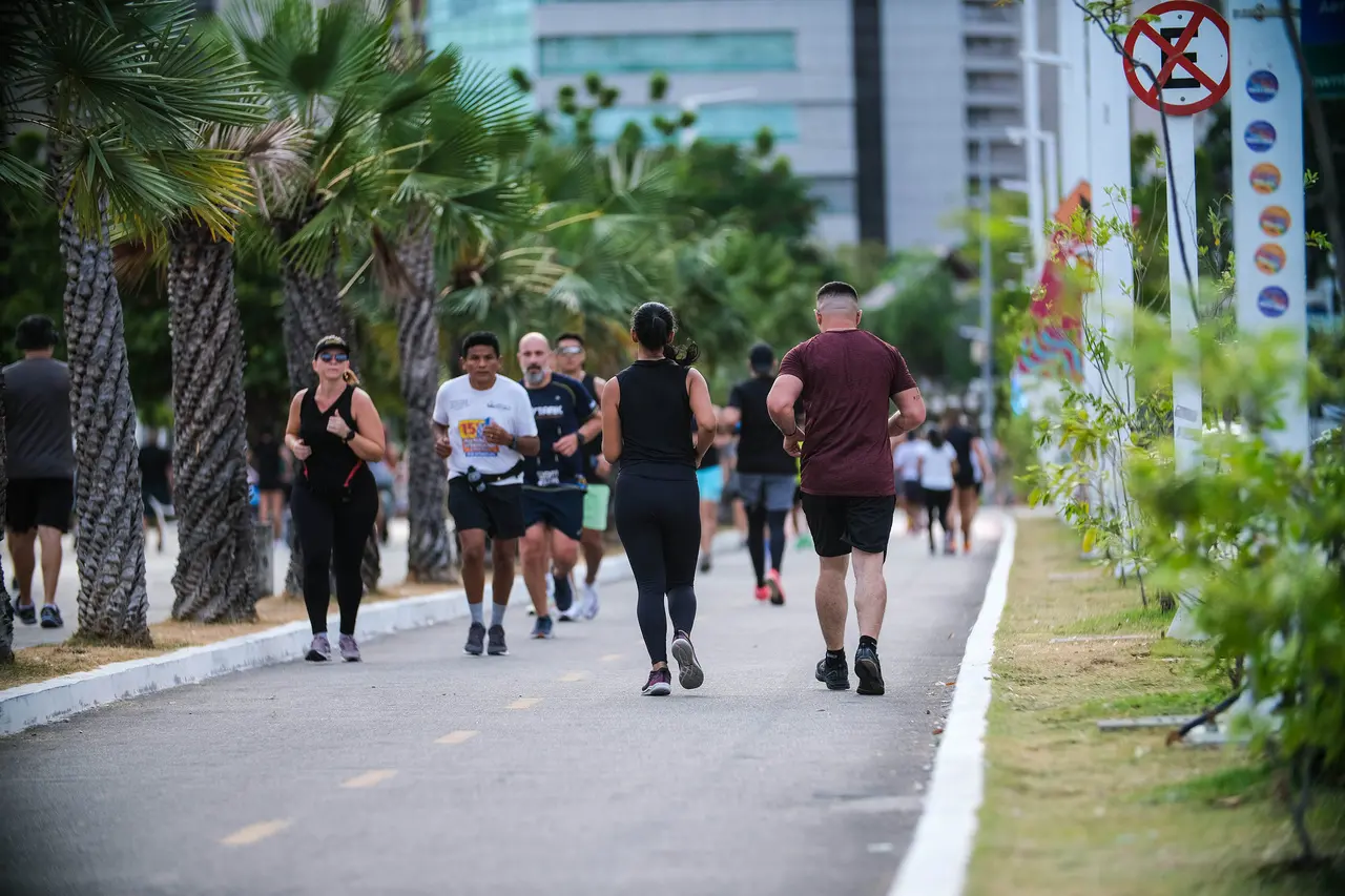 Pessoas caminhando e correndo em uma pista arborizada na orla de Fortaleza, com palmeiras ao longo do percurso, prédios ao fundo e sinalização urbana em um dia claro.