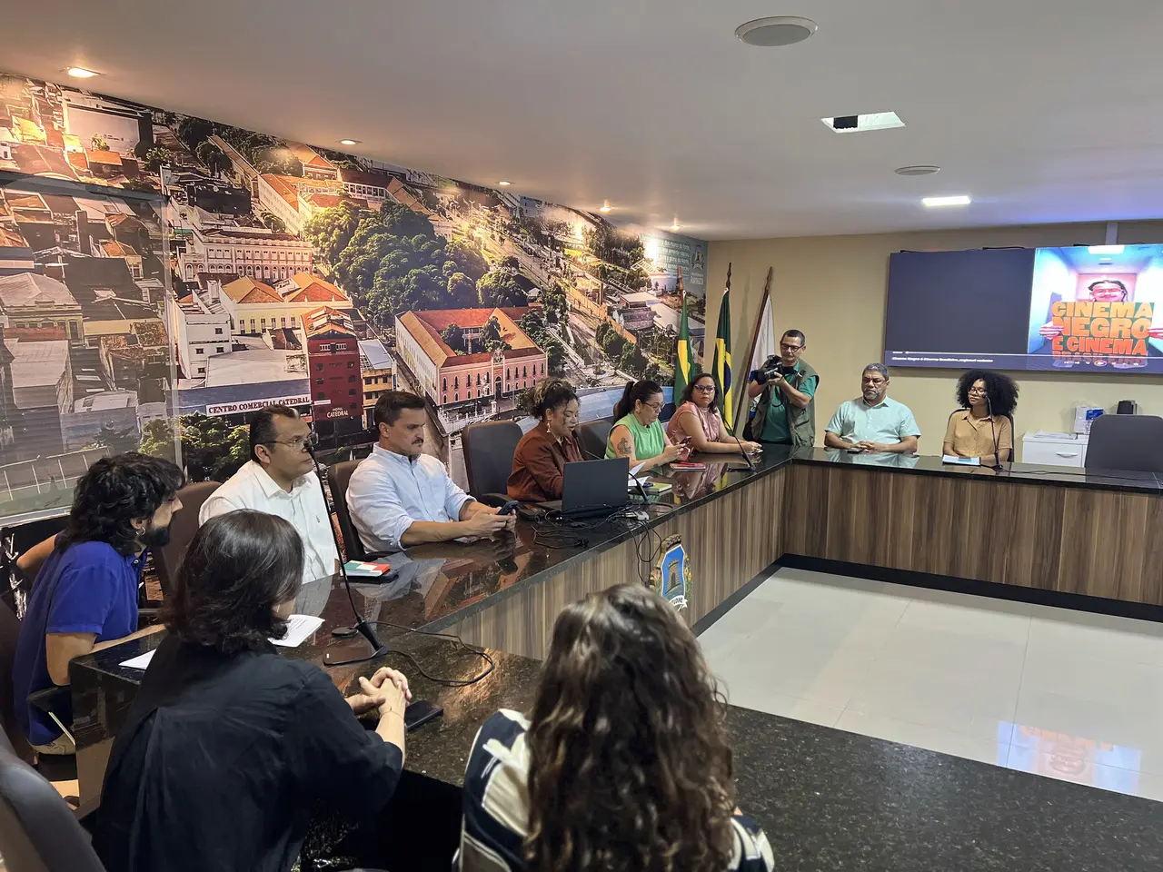 Participantes de uma audiência pública estão reunidos em torno de uma grande mesa em formato de 