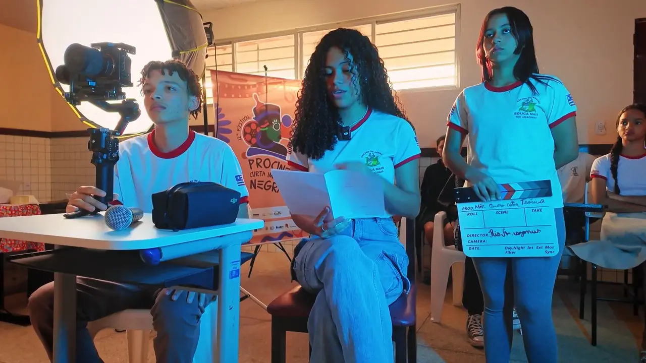 Estudantes em uniformes escolares participam de uma produção audiovisual em sala de aula, com uma jovem ao centro lendo um roteiro enquanto outra segura uma claquete ao seu lado. Equipamentos de filmagem, como uma câmera em um estabilizador e um refletor de luz, estão posicionados à esquerda, compondo o cenário da atividade prática de cinema.