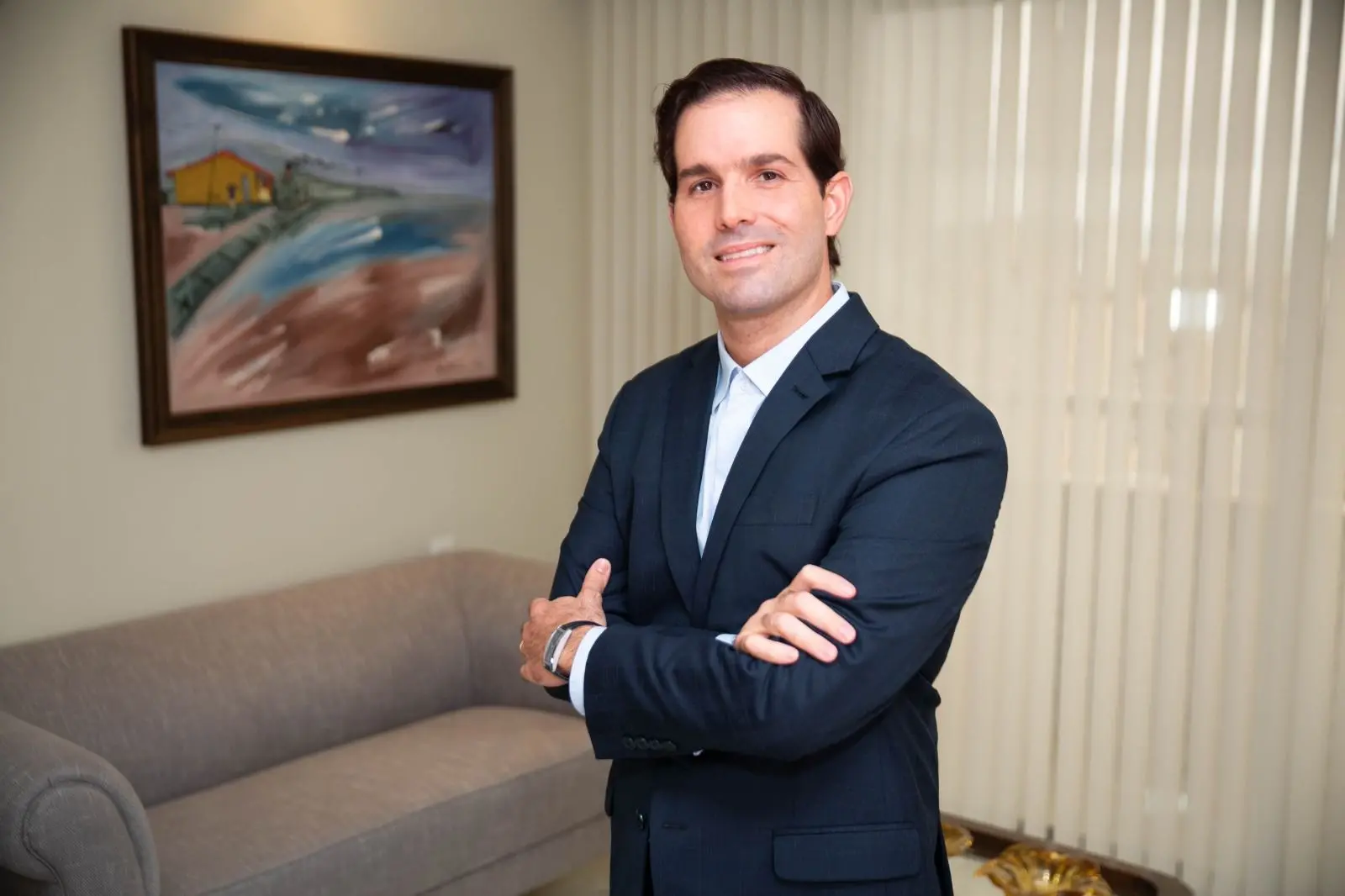 Felipe Queiroz, Homem de terno escuro posa com braços cruzados em escritório, com sofá e quadro ao fundo.