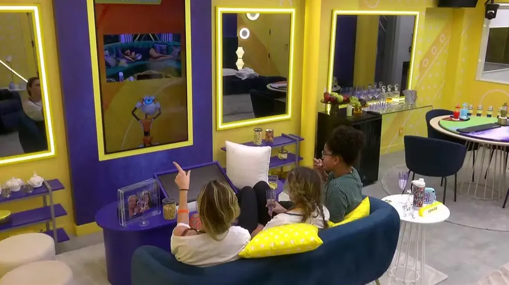 Na imagem, três participantes do BBB26 estão sentadas de costas em um sofá azul escuro, observando um grande monitor vertical na parede. A sala tem uma decoração vibrante em tons de amarelo e roxo, com espelhos retangulares iluminados por LEDs. No monitor, aparece a imagem de outra pessoa dormindo em um quarto e uma ilustração do robô 