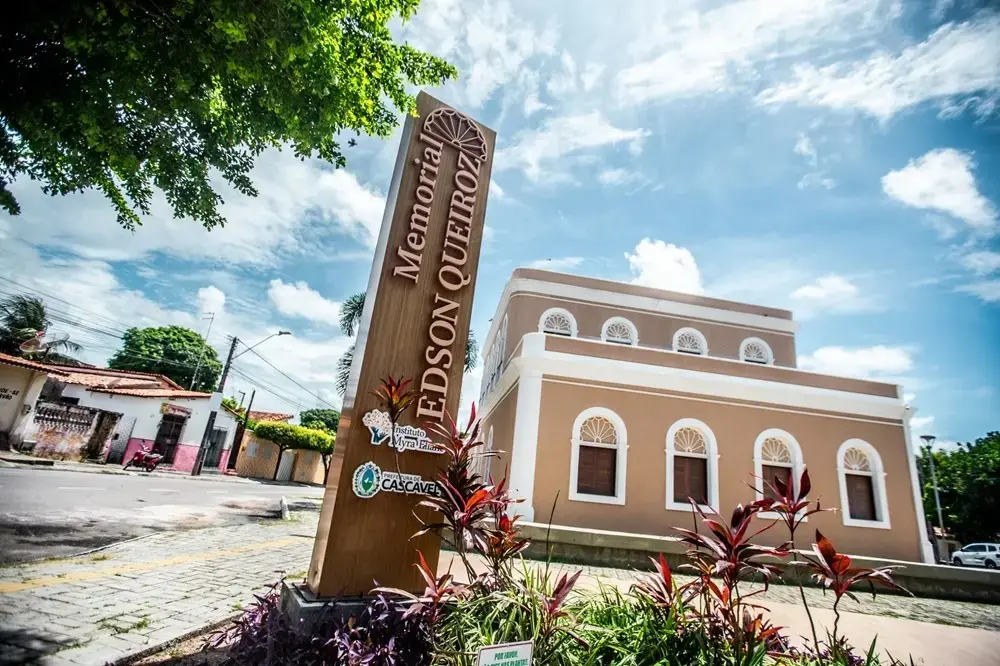 Na imagem, fotografia externa em ângulo baixo do Memorial Edson Queiroz, em Cascavel. No primeiro plano, à esquerda, destaca-se um totem vertical de madeira com a inscrição 