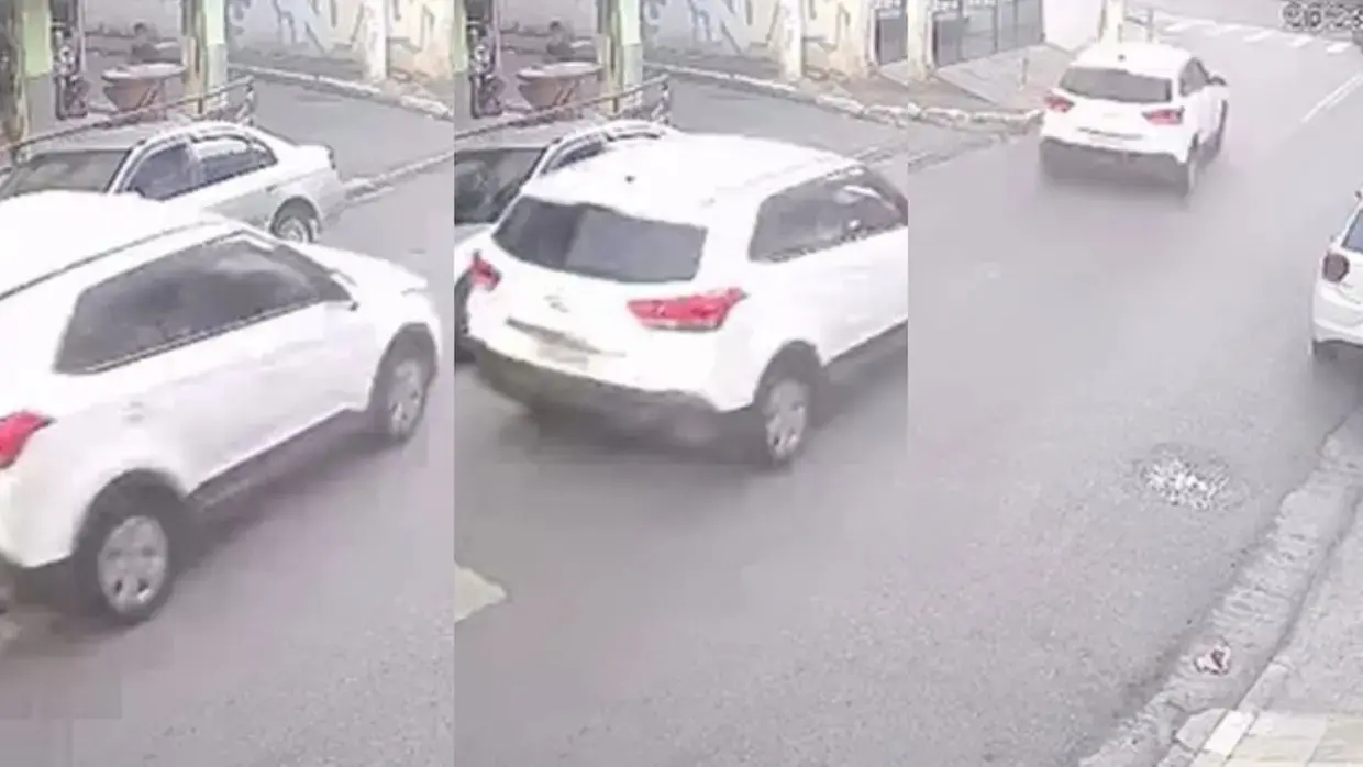 Na imagem, montagem horizontal com três capturas de tela sequenciais de uma câmera de segurança, mostrando o movimento de um SUV branco em uma rua urbana. Na primeira imagem (esquerda): O SUV branco aparece entrando no quadro, passando por um carro prata estacionado. Na segunda imagem (centro): O SUV é visto de costas, continuando seu trajeto pela via. Na terceira imagem (direita): O veículo aparece mais distante, seguindo em direção a um cruzamento. As imagens possuem baixa resolução e tons acinzentados, típicos de gravações de segurança. A rua parece estar molhada ou sob luz difusa, e há outros veículos estacionados nas laterais. No canto superior direito da terceira imagem, há um pequeno selo de crédito indicando 