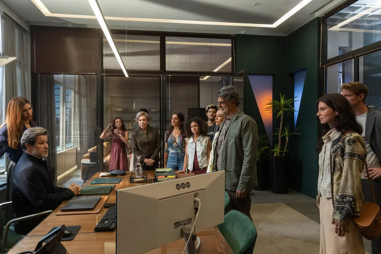 Grupo de pessoas reunidas em um escritório moderno, posicionadas em frente a uma mesa com computador, enquanto alguém sentado parece conduzir uma conversa ou reunião; o ambiente tem janelas amplas, iluminação linear no teto e plantas decorativas.