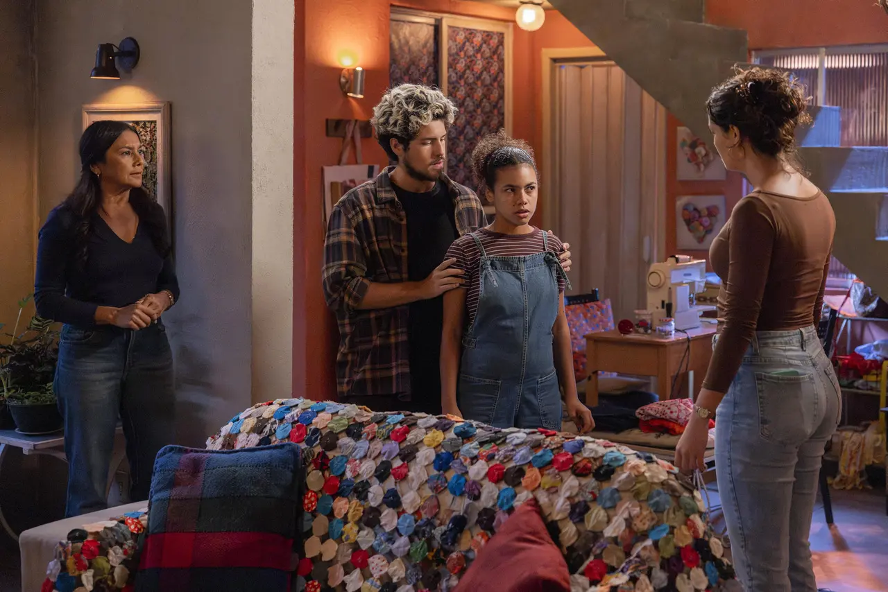 Quatro pessoas estão reunidas em uma sala aconchegante, decorada com colchas coloridas, máquina de costura e iluminação quente; duas permanecem lado a lado no centro enquanto outras observam, criando uma cena de tensão e expectativa.