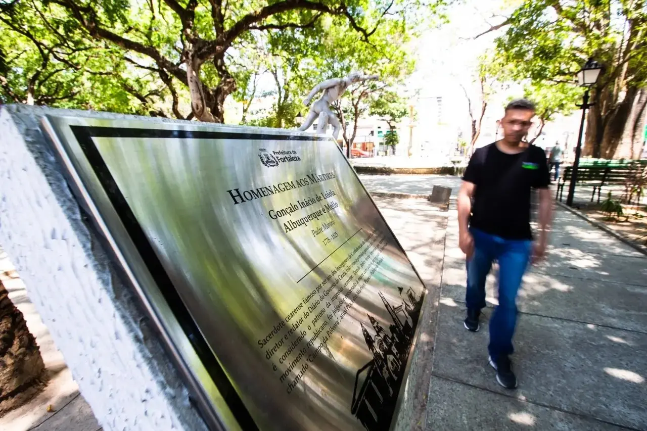 Placa com homenagem a mártires instalada no Passeio Público. Ao lado, um pedestre caminha no local.
