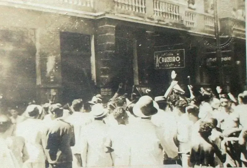 Multidão reunida em frente a um prédio histórico, em cena antiga em preto e branco. Várias pessoas, muitas usando chapéus, observam ou participam de uma aglomeração diante de uma fachada com sacada e uma placa com o nome “A Cruzeiro”. A imagem tem aparência desgastada, típica de fotografia histórica, e sugere um evento público ou manifestação.