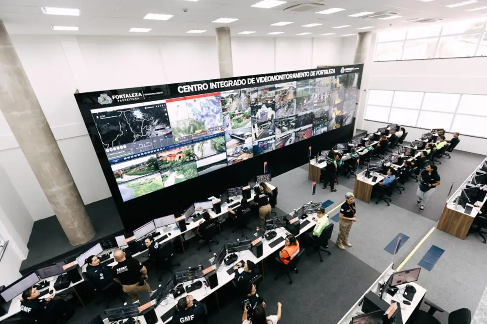 Na imagem, uma visão aérea de ângulo aberto mostra o interior do Centro Integrado de Videomonitoramento de Fortaleza. No centro da sala, há um imenso painel de vídeo (videowall) inclinado, exibindo dezenas de telas com imagens de câmeras de segurança, mapas e gráficos de monitoramento urbano. No topo do painel, destaca-se o logotipo da Prefeitura de Fortaleza e o nome do centro. Diversos operadores e agentes de segurança, vestindo uniformes de diferentes corporações (como a Guarda Municipal - GMF), estão posicionados em fileiras de mesas organizadas à frente e ao lado do painel. Eles operam computadores individuais com múltiplos monitores. O ambiente é amplo, moderno, com pé-direito alto, colunas de concreto aparente, iluminação clara e carpete cinza no chão. Agentes circulam pelo espaço, indicando um ambiente de trabalho ativo e coordenado.