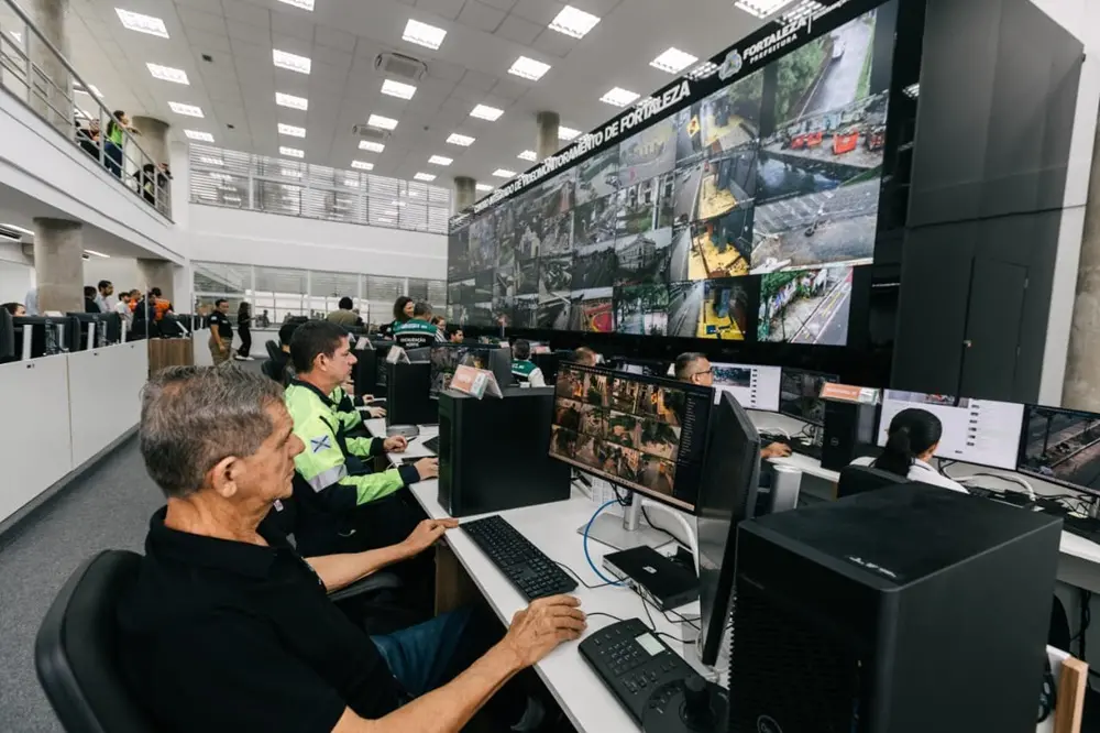Na imagem, fotografia em ângulo lateral e de média distância do Centro Integrado de Videomonitoramento de Fortaleza. Em primeiro plano, um homem de perfil, vestindo camisa polo preta, opera um computador com as mãos no teclado e no mouse. Ao lado dele, outros agentes de segurança, incluindo um homem com uniforme verde-limão e azul, trabalham em suas respectivas estações. As mesas brancas estão equipadas com monitores que exibem múltiplos mosaicos de câmeras de segurança e teclados de controle específicos para vigilância. Ao fundo, à direita, destaca-se o grande painel de videowall preto com o nome do Centro e o brasão da Prefeitura de Fortaleza, exibindo diversas imagens em tempo real de vias públicas. O ambiente possui arquitetura moderna com mezanino à esquerda, onde outras pessoas podem ser vistas, e colunas de concreto aparente sob uma iluminação intensa de painéis de LED no teto.
