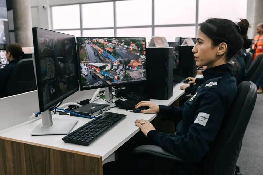 Na imagem, uma mulher, vestindo um uniforme azul-escuro com o brasão da Prefeitura de Fortaleza em uma das mangas, está sentada à frente de dois monitores em um escritório. Ela possui o cabelo preso e está segurando o mouse em sua mão direita. No monitor à direita, há quatro imagens de câmeras de segurança ao vivo. No monitor à esquerda, há seis. O escritório possui uma parede branca e uma janela ao fundo.