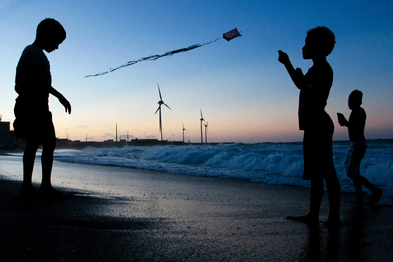 Crianças brincam na areia da praia ao entardecer, com suas silhuetas destacadas contra o céu suave enquanto uma delas empina uma pipa. Ao fundo, as ondas quebram na orla e turbinas eólicas se alinham no horizonte, completando a paisagem litorânea tranquila.