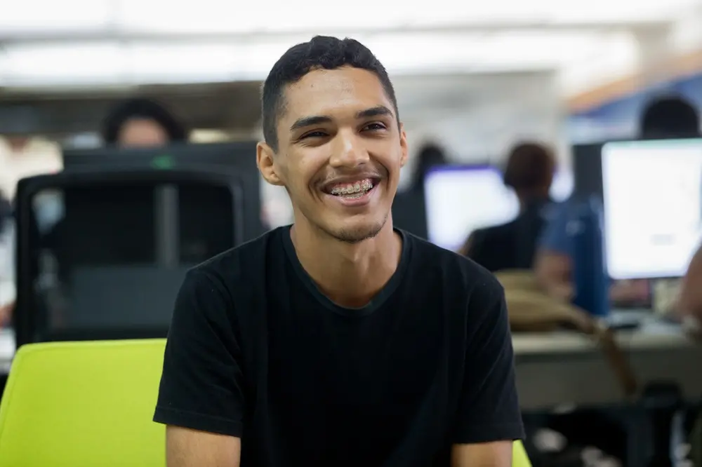 Na imagem, fotografia de meio corpo de um homem jovem de pele parda, cabelos curtos e camiseta preta. Ele está sentado em uma cadeira de escritório verde-limão e olha para o lado com um sorriso largo, exibindo o aparelho ortodôntico. O ambiente é um escritório moderno e iluminado, com outras pessoas e monitores de computador desfocados ao fundo, criando uma profundidade de campo que destaca o rapaz em primeiro plano.
