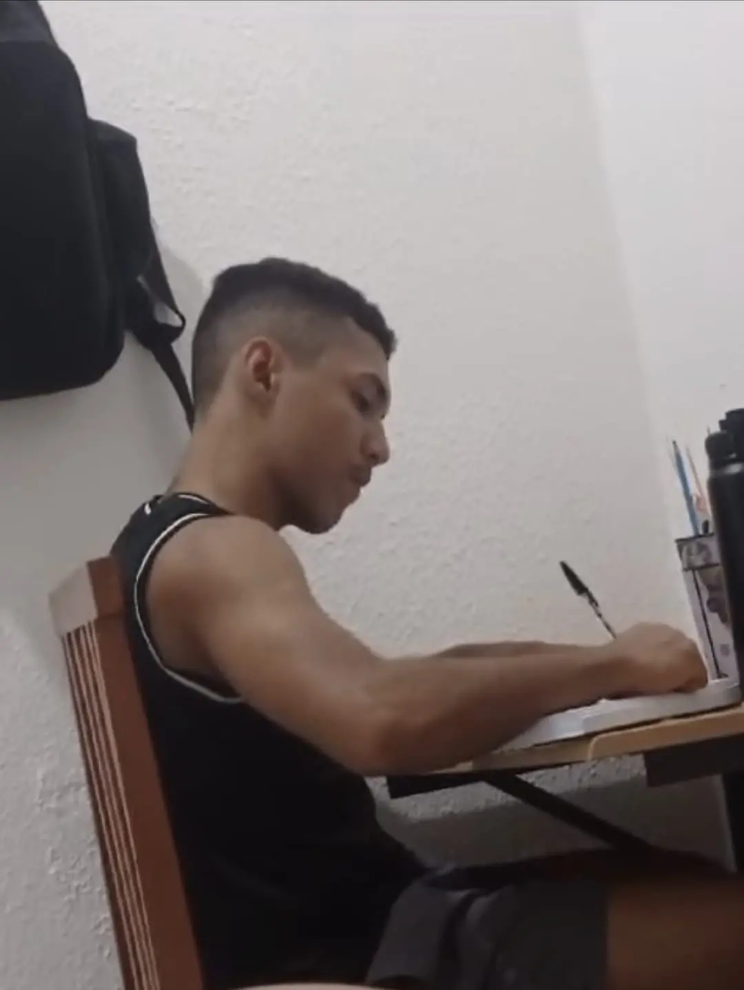 Na imagem, fotografia de perfil de um jovem de pele parda e cabelos curtos com as laterais raspadas. Ele está sentado em uma cadeira de madeira em frente a uma mesa, concentrado enquanto escreve em um caderno com uma caneta. O rapaz veste uma camiseta regata preta. Ao fundo, vê-se uma parede branca com textura leve e uma mochila pendurada no canto superior esquerdo. Sobre a mesa, há alguns objetos de escritório e uma garrafa preta. A iluminação vem de cima, criando sombras suaves.
