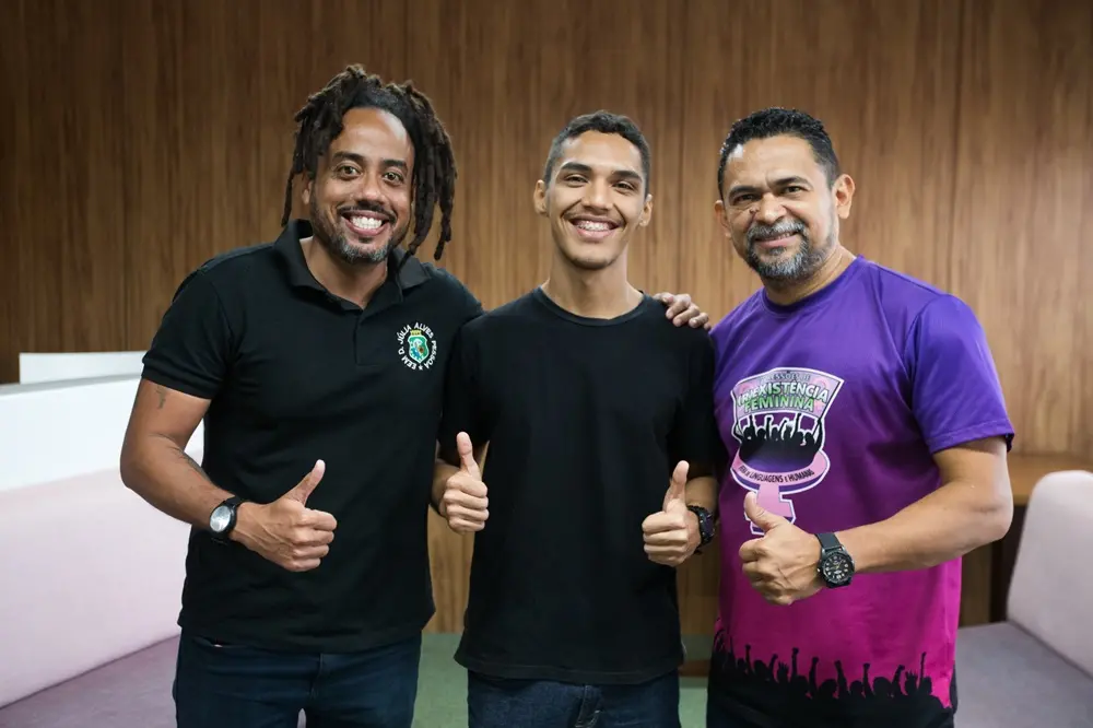 Na imagem, três homens sorridentes posam para uma foto em um ambiente interno. No centro, um jovem de pele parda, cabelos curtos e camiseta preta faz um sinal de 