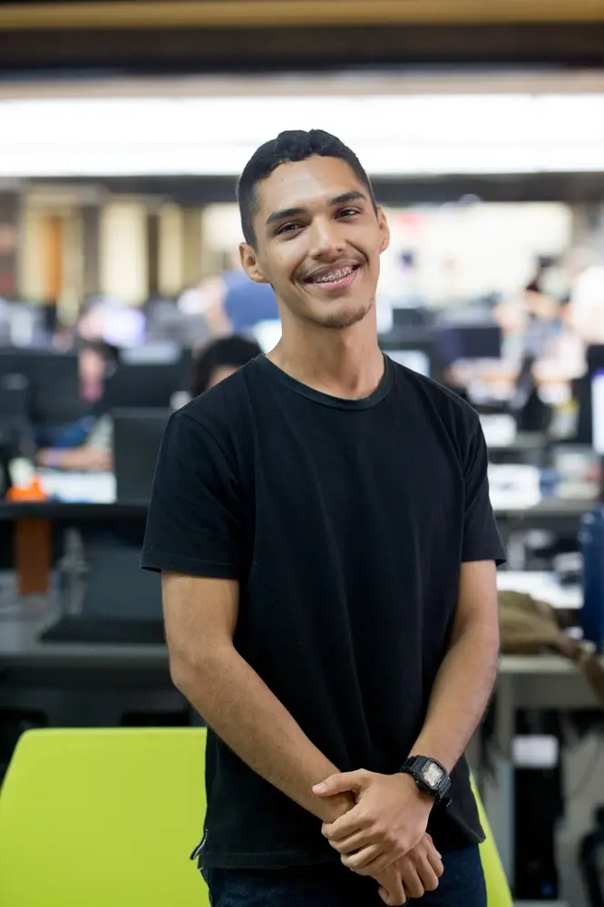 Na imagem, fotografia de plano médio de um jovem de pele parda, cabelos pretos curtos e camiseta preta. Ele está de pé, sorrindo para a câmera e exibe aparelho ortodôntico. Suas mãos estão cruzadas à frente do corpo e ele usa um relógio digital preto no pulso esquerdo. O homem está posicionado à frente de uma cadeira de escritório verde-limão. O fundo mostra um ambiente de escritório amplo e iluminado, com mesas e computadores propositalmente desfocados para destacar a pessoa em primeiro plano.