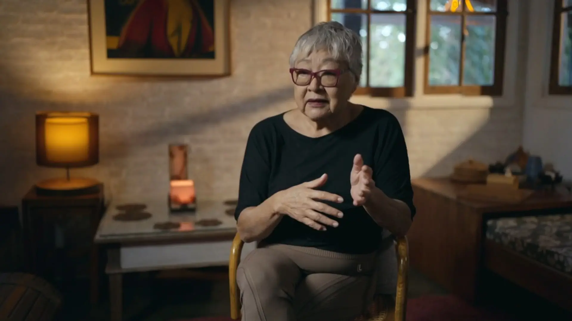 A cineasta Tizuka Yamasaki aparece sentada em uma cadeira de palha, gesticulando com as mãos enquanto concede uma entrevista para a série 'O Testamento - O Segredo de Anita Harley'. Ela veste uma blusa preta, óculos de armação vermelha e está posicionada diante de uma parede de tijolos aparentes iluminada por um abajur de luz quente ao fundo.