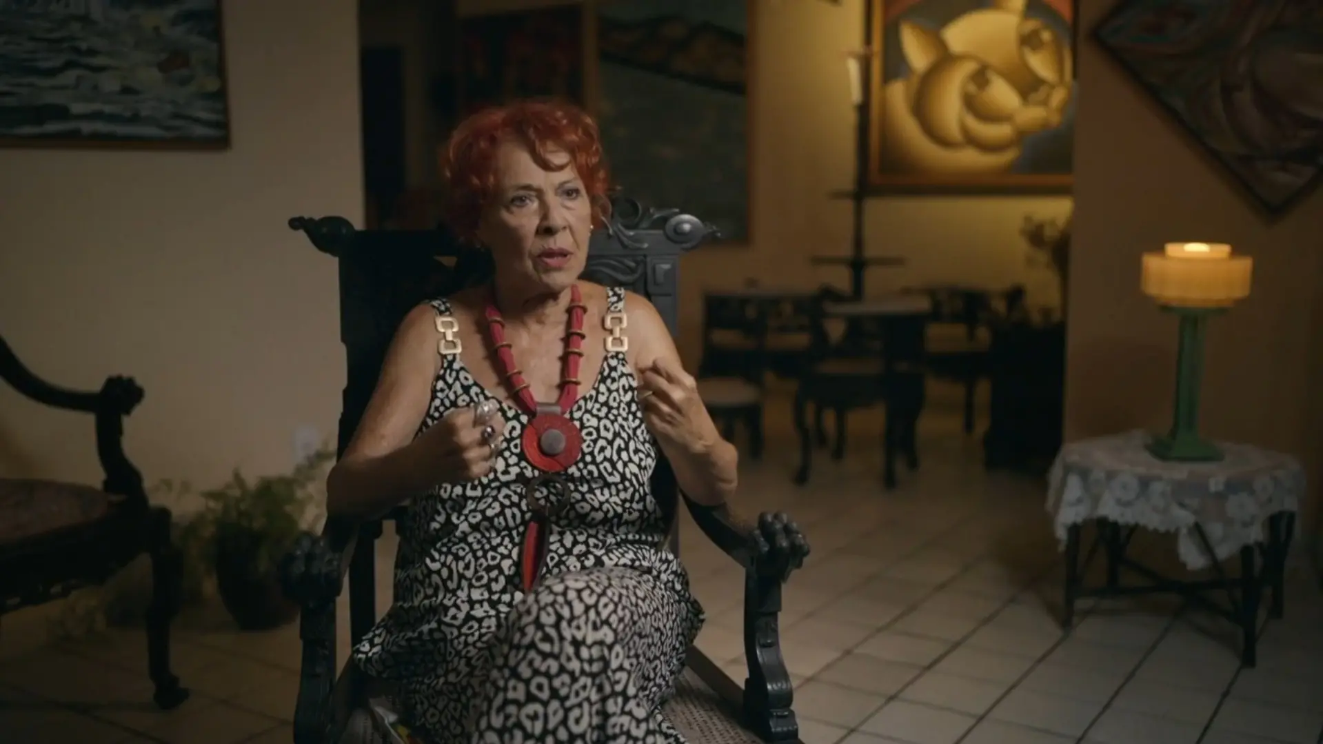 A cineasta Kátia Mesel aparece sentada em uma cadeira de madeira entalhada, gesticulando com as mãos durante entrevista para a série 'O Testamento - O Segredo de Anita Harley'. Ela veste um macacão com estampa de animal print e um colar vermelho marcante, em um ambiente decorado com quadros e uma iluminação suave de abajur ao fundo.