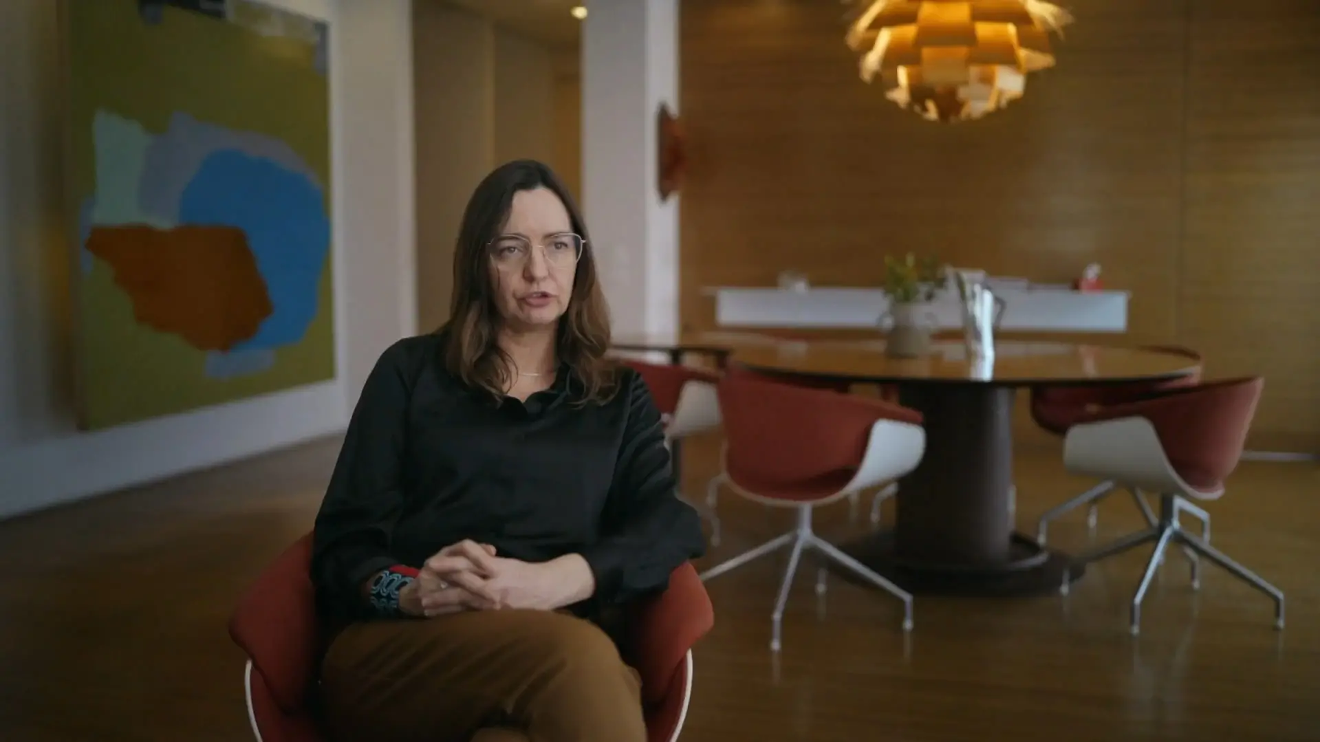 A montadora Karen Harley aparece sentada em uma cadeira moderna de cor terracota, com as mãos entrelaçadas durante entrevista para a série 'O Testamento - O Segredo de Anita Harley'. Ela veste uma blusa preta, calça marrom e óculos, posicionada em uma sala elegante que exibe uma tela abstrata colorida na parede e uma luminária de design escultural ao fundo.