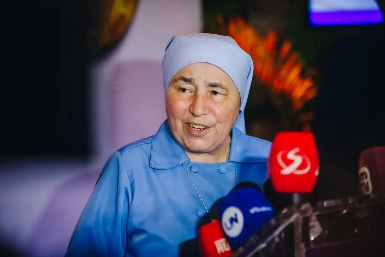 Mulher idosa, de pele clara, veste hábito religioso azul claro, com véu cobrindo a cabeça e roupa de botões frontais. Ela fala com a imprensa, olhando levemente para o lado, diante de vários microfones com logotipos de emissoras. O ambiente é interno, com iluminação quente e fundo desfocado com elementos decorativos.
