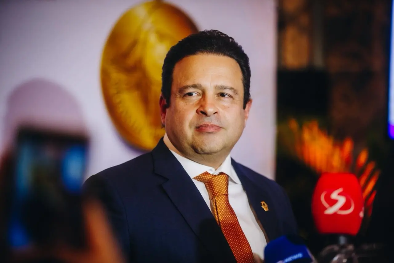 Homem de meia-idade, de pele clara e cabelos escuros, veste terno azul escuro, camisa branca e gravata laranja. Ele fala com a imprensa, olhando levemente para o lado, enquanto microfones com logotipos de emissoras aparecem em primeiro plano. Ao fundo, há um ambiente interno com iluminação quente e elementos decorativos, incluindo um objeto dourado desfocado na parede.