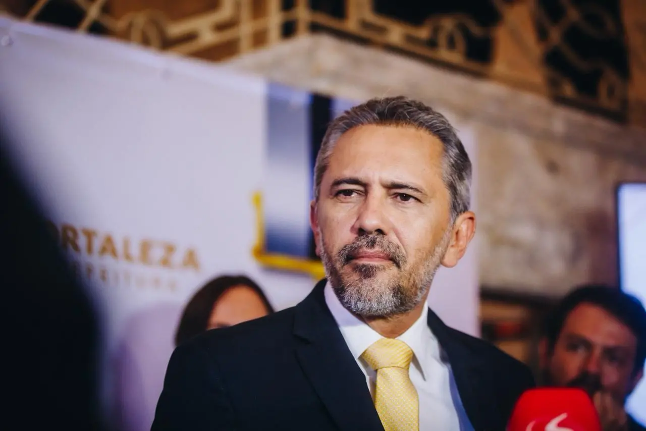 Homem de meia-idade, com cabelos grisalhos e barba aparada, veste terno escuro, camisa branca e gravata amarela. Ele está em primeiro plano, com expressão séria e olhar direcionado para a lateral. Ao fundo, desfocado, aparecem outras pessoas e um painel com a palavra “Fortaleza”, indicando um ambiente institucional ou evento oficial. Um microfone vermelho surge parcialmente na parte inferior direita da imagem.