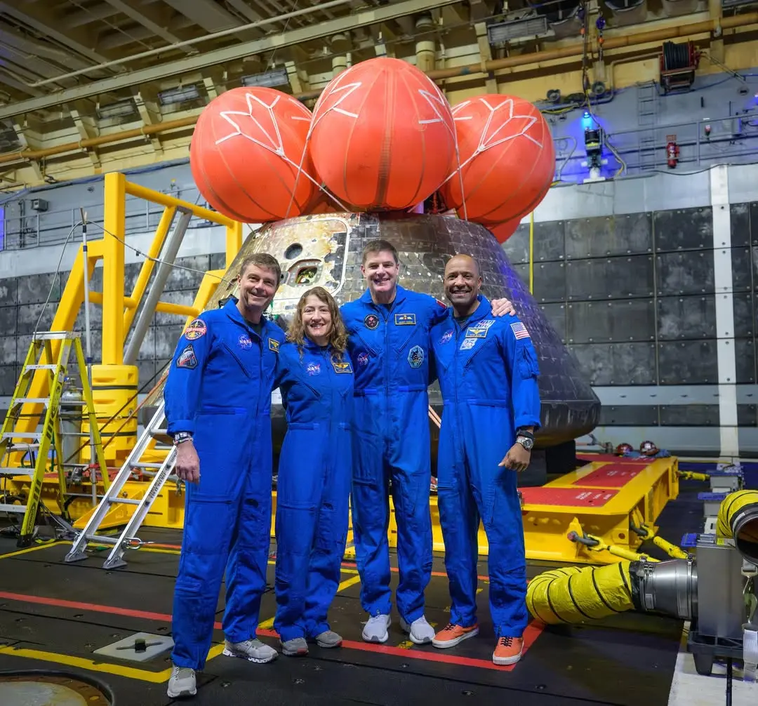 Os quatro astronautas da missão Artemis II sorriem abraçados à frente da cápsula Orion, vestindo macacões azuis da NASA em um hangar de recuperação. A nave exibe marcas de reentrada atmosférica.