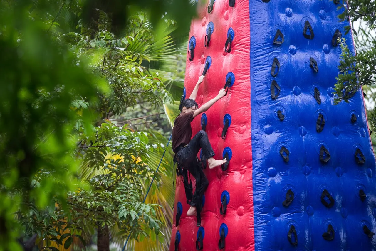 Pessoa escalando uma parede de escalada inflável vermelha e azul ao ar livre, cercada por vegetação tropical, usando as mãos e os pés enquanto está presa a um equipamento de segurança.