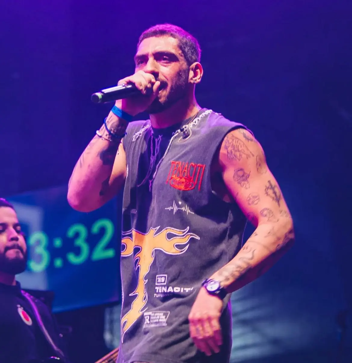 Cantor se apresenta no palco segurando um microfone, sob iluminação roxa, vestindo camiseta sem mangas com estampa e exibindo tatuagens nos braços, enquanto outro músico aparece ao fundo.