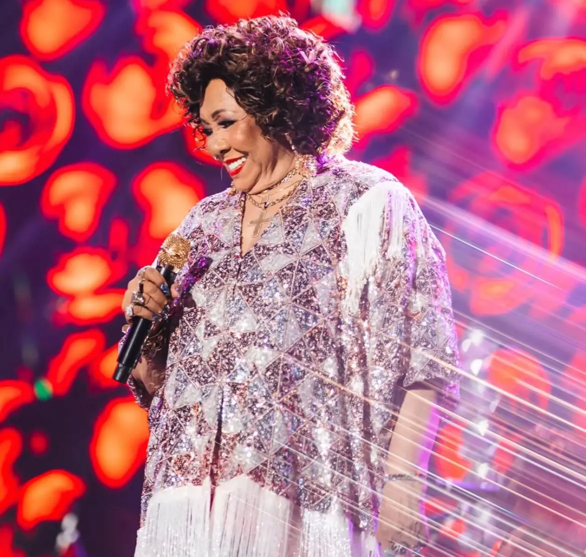 Cantora se apresenta no palco sorrindo, segurando um microfone, vestindo roupa brilhante com franjas e acessórios, sob iluminação intensa e fundo com luzes vermelhas desfocadas.
