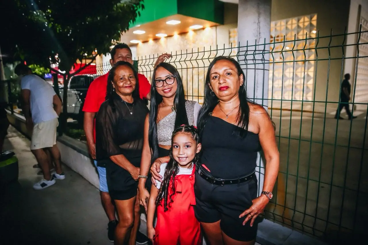 Grupo de cinco pessoas posa para foto em área externa à noite, incluindo três mulheres, um homem ao fundo e uma criança à frente usando roupa vermelha, ao lado de uma grade e de um prédio iluminado.