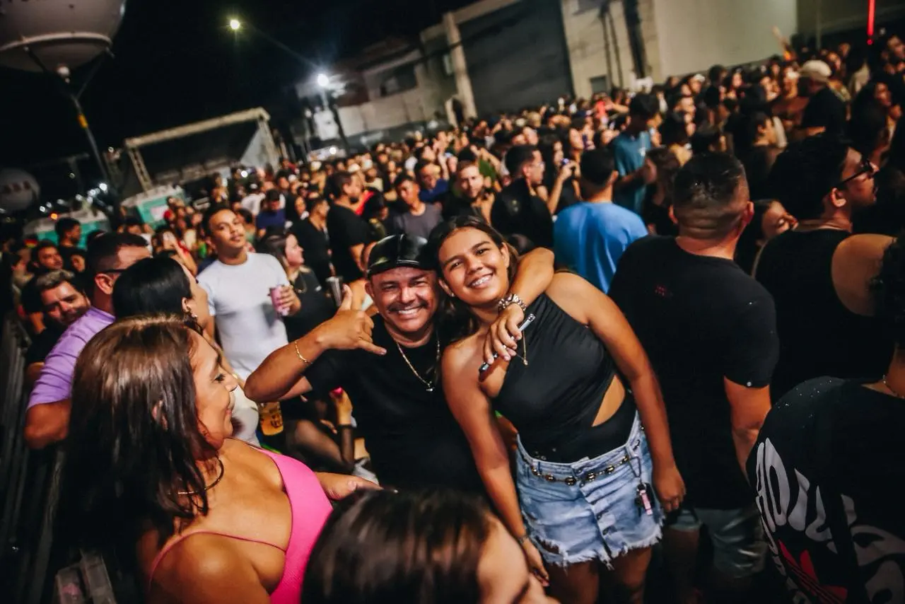 Uma mulher com um top preto e shorts jeans sorri abraçando um homem de boné preto que faz um sinal de 