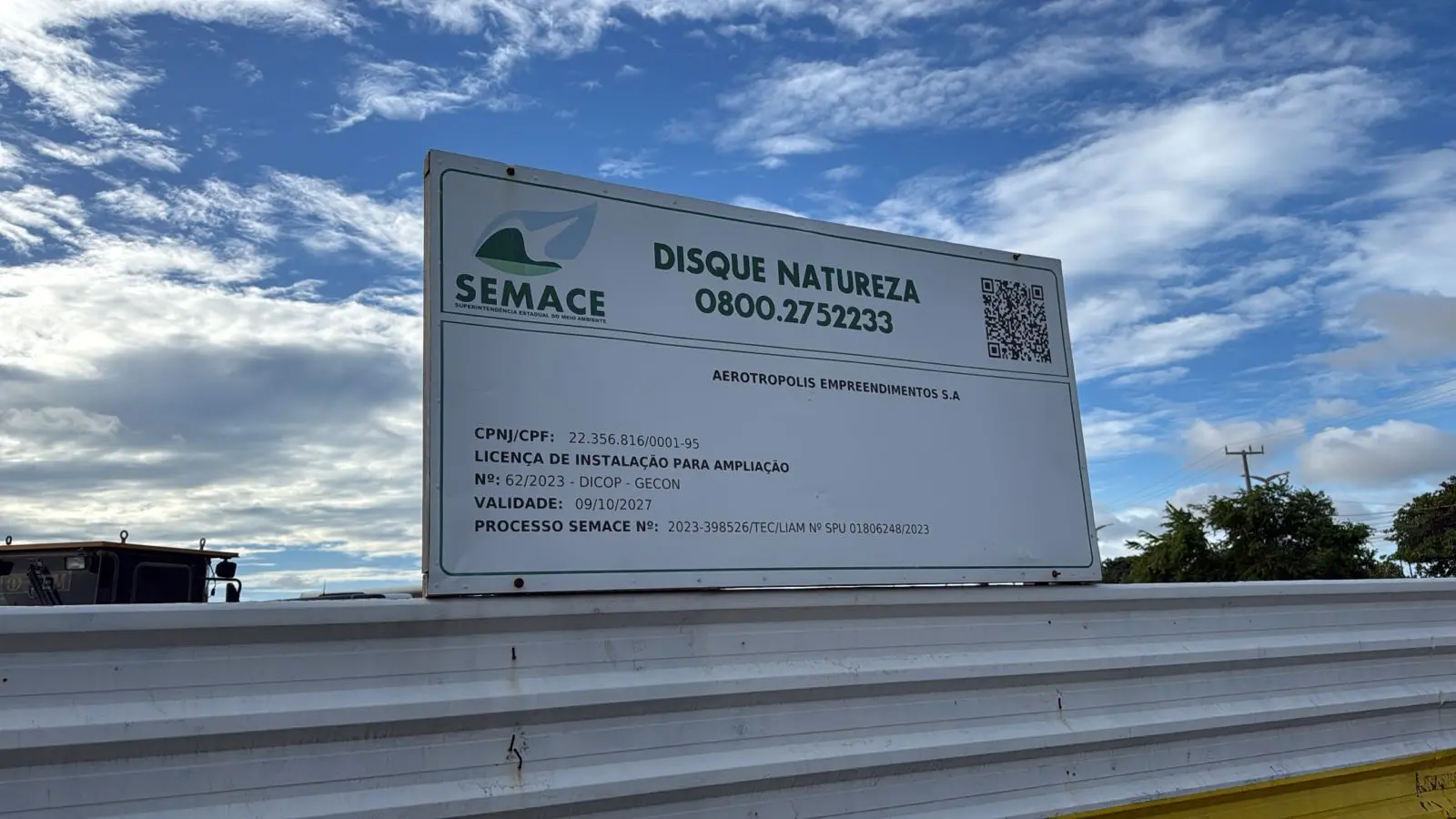 Placa informativa da SEMACE instalada sobre um tapume metálico branco, em área externa, sob céu azul com nuvens. A placa exibe o texto “DISQUE NATUREZA 0800.2752233”, nome Aeroópolis Empreendimentos S.A., dados de licença de instalação para ampliação (com validade até 09/10/2027), número de processo da Semace e um QR code no canto superior direito.