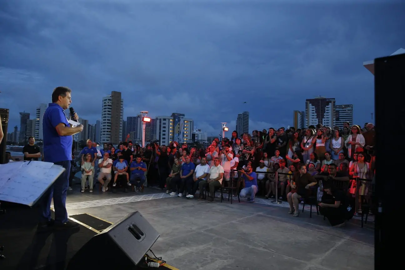 Prefeito discursa em palco ao ar livre durante evento de aniversário de Fortaleza, com público reunido ao entardecer.