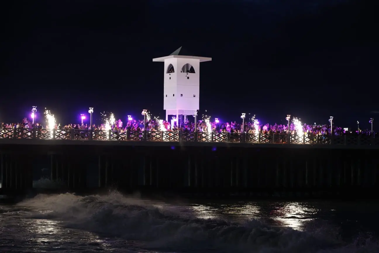 Ponte metálica vista de longe à noite com luzes roxas e fogos.