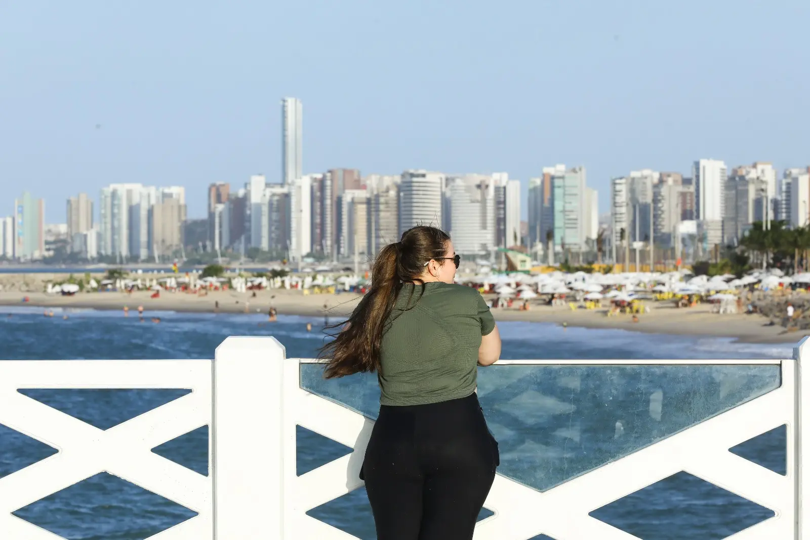 Pessoa de costas, com cabelo preso e usando óculos escuros, apoia-se em um parapeito branco e observa o mar. Ao fundo, há uma praia movimentada com guarda-sóis e uma linha de prédios altos ao longo da orla sob céu azul.